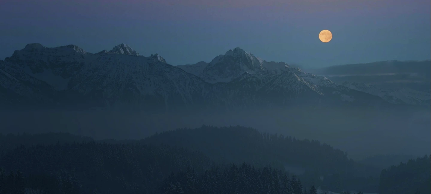 A full moon rising over mountain peaks under a dark sky